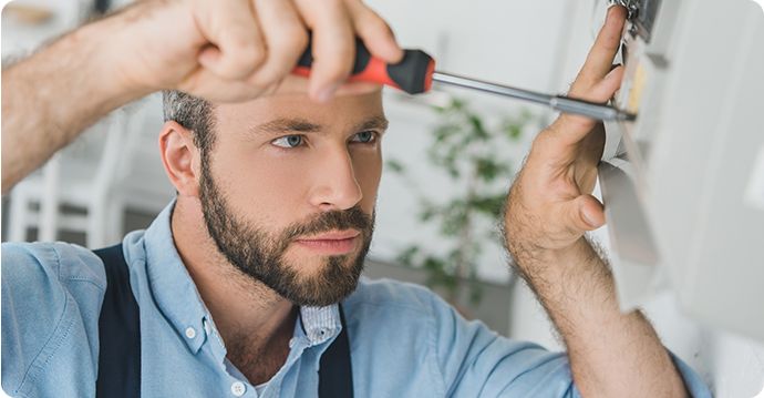 Osoba z brodą i w niebieskiej koszuli używa śrubokręta, aby dopasować element wyposażenia na ścianie. W rozmytym tle widać roślinę, co wskazuje na ich dbałość o szczegóły, niezależnie od tego, czy chodzi o instalację klimatyzacji Gree, czy o wykonanie serwisu gwarancyjnego.