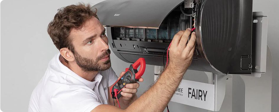 Mężczyzna skrupulatnie sprawdza okablowanie ściennej jednostki klimatyzacyjnej za pomocą miernika cęgowego. Ubrany w białą koszulę, wydaje się być całkowicie skupiony na swoim zadaniu.