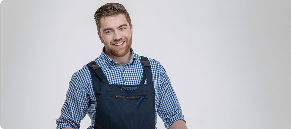 Uśmiechnięty mężczyzna z brodą, wyglądający na gotowego do całodniowego serwisu pogwarancyjnego, ubrany w niebieską kraciastą koszulę i ciemny kombinezon, stoi na jednolitym jasnoszarym tle.