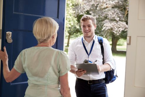 Młody mężczyzna z notatnikiem i smyczą stoi przy niebieskich drzwiach wejściowych, rozmawiając o Klimatyzacja Gree ze starszą kobietą. Ma na sobie jasnozielony top, cieszy się słonecznym dniem z drzewami w tle, podczas gdy oni zagłębiają się w szczegóły opcji instalacji.